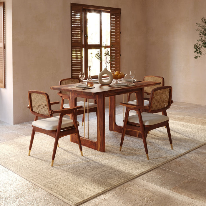 A mid-century modern dining set featuring a rectangular wooden table and four matching armchairs with woven cane backs and light beige upholstered seats, styled on a textured rug in a warm, neutral-toned room.